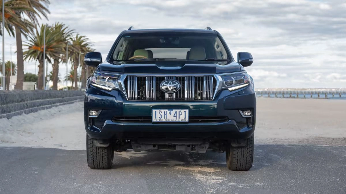 Dark blue Toyota Land Cruiser Prado SUV front view parked by ocean with palm trees and cloudy sky