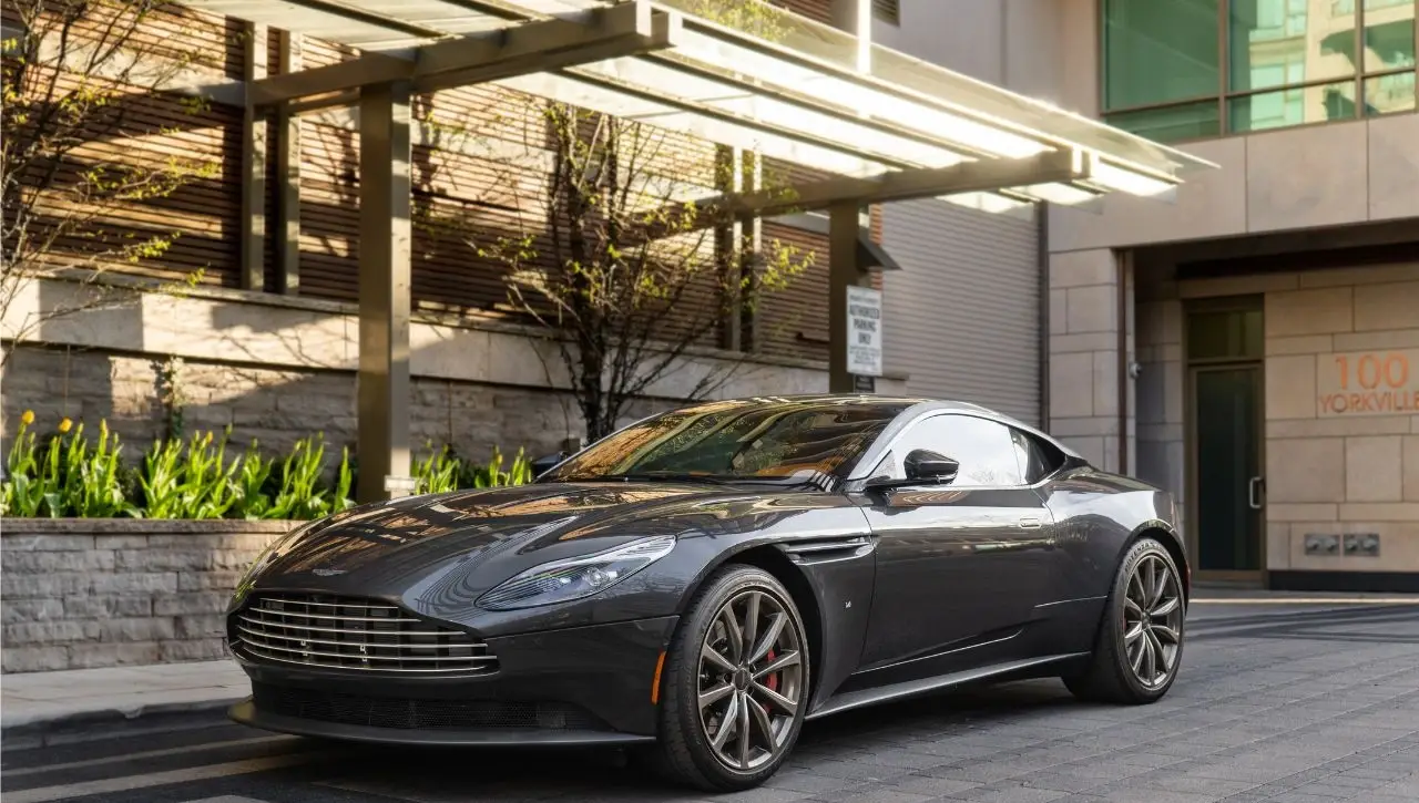 Black Aston Martin DB12 sports car parked under architectural pergola with modern building and landscaping