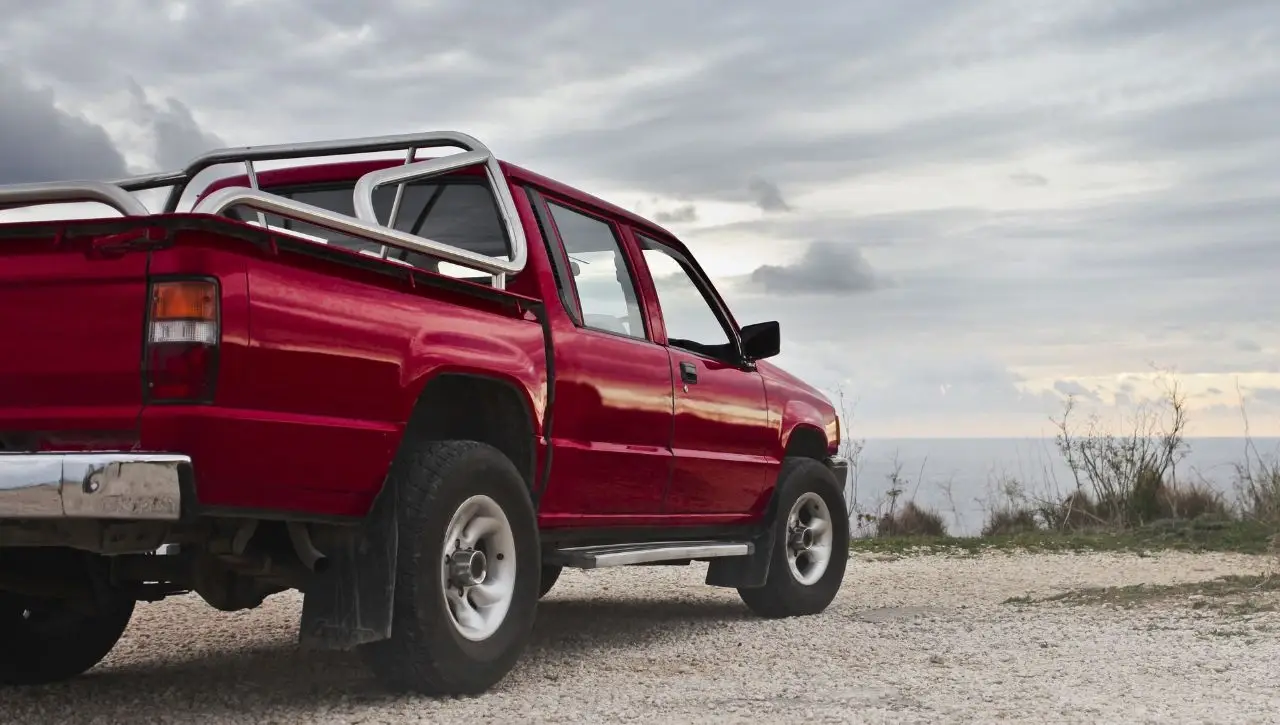 Red pickup truck with roll bar parked on sandy terrain with cloudy sky background