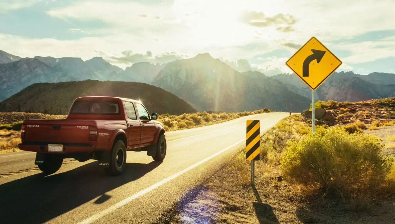 Red pickup truck driving on winding mountain highway with yellow turn warning sign and scenic landscape
