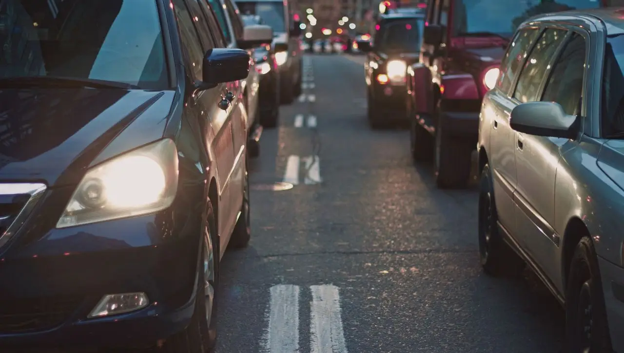 Multiple American muscle cars in traffic on city street at dusk with car headlights and taillights illuminated