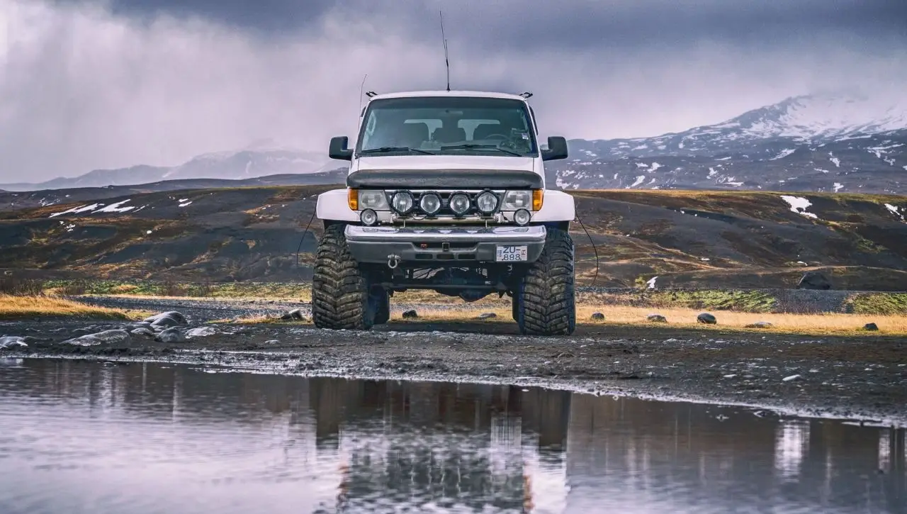 White Toyota Land Cruiser LC76 Station Wagon with roof rack parked in shallow water with snow-capped mountains in background