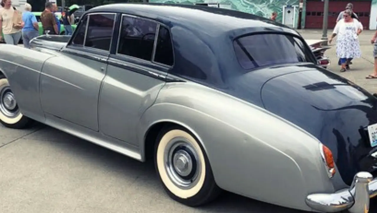 Two-tone silver and dark blue Bentley S Series classic sedan parked at outdoor restoration event with mountains in background